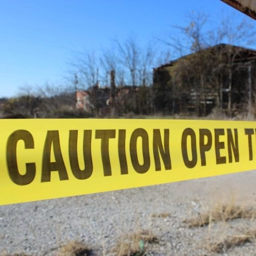 Yellow barricade tape that reads "CAUTION OPEN" before being cut off. The background has a few large, blurry buildings obstructed by trees.